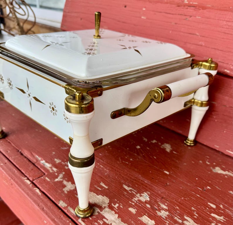 May include: White and gold vintage serving dish with a glass lid. The dish has gold accents, a handle, and decorative starburst and floral patterns. The legs and handles are white with gold trim. A gold knob is on the lid.