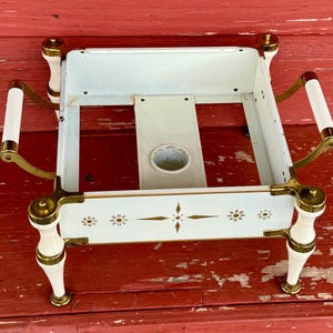 May include: Vintage white metal serving tray with brass accents and decorative floral designs. Features white handles and legs with brass detailing. The tray has a central opening and is set against a red wooden surface.