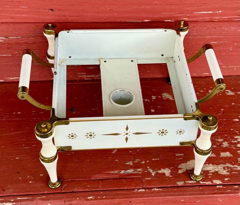 May include: Vintage white metal serving tray with brass accents and decorative floral designs. Features white handles and legs with brass detailing. The tray has a central opening and is set against a red wooden surface.