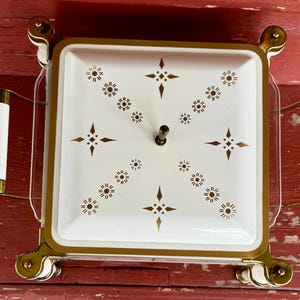 May include: A vintage square serving dish with a white lid featuring a gold starburst pattern. The dish has gold trim and handles with white accents. The dish sits on a gold metal stand.