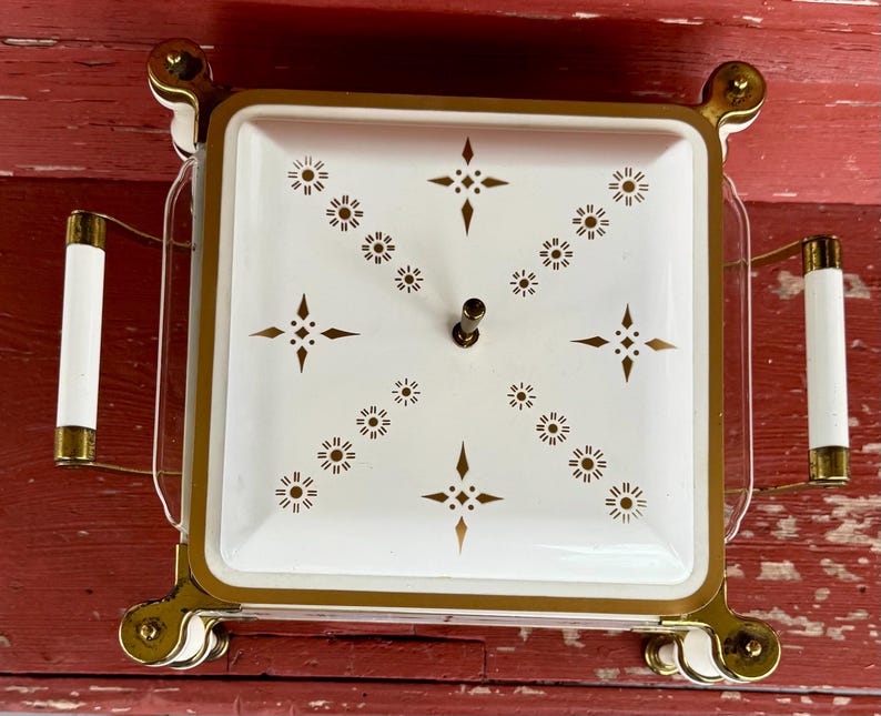May include: A vintage square serving dish with a white lid featuring a gold starburst pattern. The dish has gold trim and handles with white accents. The dish sits on a gold metal stand.