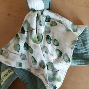 May include: A soft baby security blanket featuring a white fabric top with a green eucalyptus leaf pattern and a solid sage green fabric underside. The blanket has a central knotted head section and a small fabric tag labelled with the text aya made.