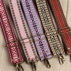 May include: Five colorful woven bag straps with geometric patterns. The straps feature a variety of colors, including red, pink, purple, brown, and a mix of other hues. Each strap has metal hardware for attachment.