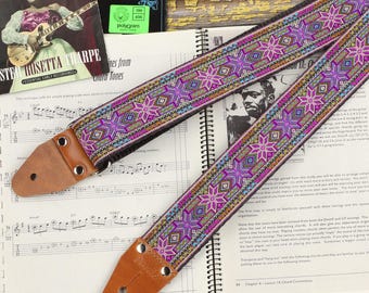 May include: A colorful guitar strap with a pink, purple, and blue geometric pattern. The strap has a brown leather end with metal eyelets.