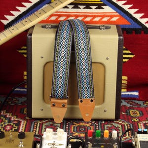Può includere: Due cinturini per chitarra con sfondo nero e motivi geometrici colorati. I cinturini sono realizzati in tessuto intrecciato e hanno estremità in pelle marrone.