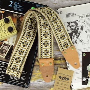 May include: A tan and brown guitar strap with a geometric pattern. The strap is made of woven fabric and has a leather end with a brass buckle. The strap is labeled "Pardo Guitar Straps" and "PGS Artisan Handmade Straps".