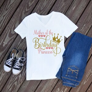 May include: White t-shirt with a gold glitter design that says "Mother of the Birthday Princess" with a gold crown and pink hearts.