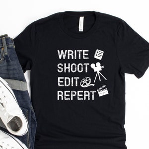 Könnte beinhalten: Schwarzes T-Shirt mit weißem Text "WRITE SHOOT EDIT REPEAT" und filmbezogenen Symbolen. Das Shirt wird auf weißem Hintergrund mit einer blauen Jeans und weißen Turnschuhen gezeigt.
