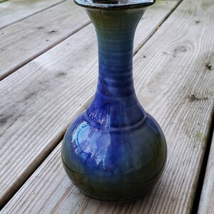 May include: A ceramic vase with a bulbous base and a slender, flared neck. The glaze features a gradient transition from a deep, vibrant blue in the center to a dark, earthy olive green at the bottom and a dark rim at the top opening.