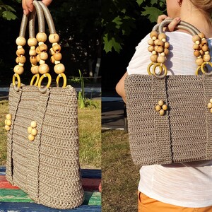 May include: A crocheted tote bag with wooden bead accents and yellow rings on the handles. The bag is a light brown color and has a textured weave.
