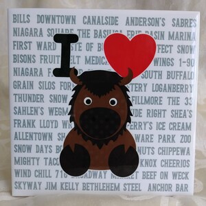 May include: A white ceramic tile with a brown and black buffalo illustration and a red heart with the text "I love" in black. The tile is covered in gray text, including the words "Bills", "Downtown", "Canalside", "Anderson's", "Sabres", "Niagara Square", "First Ward", "Bisons Fruit", "Grain Silos", "Thunder Snow", "Sahlen's", "Frank Lloyd Wright", "Allentown", "Snow Days", "Mighty Taco", "Wind Chill", "Skyway", "Jim Kelly", "Bethlehem Steel", "Anchor Bar", "Erie Basin", "Marina", "South Buffalo", "Fillmore", "The 33", "Right Shea's", "Ferry's Ice Cream", "Ware Park Zoo", "Nuts Chippewa", "Knox Cheerios", "Beef on Weck", "Broadway Market".