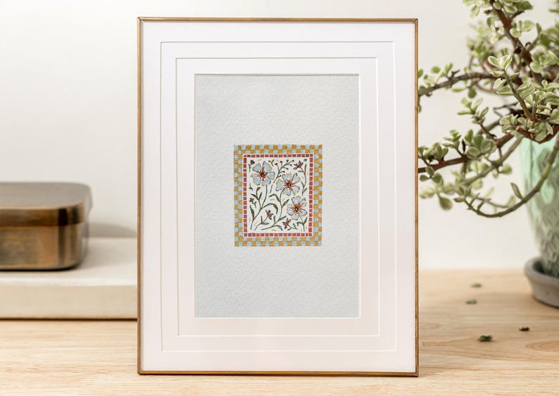 K&ouml;nnte beinhalten: Ein gerahmtes Aquarellbild mit einem floralen Design mit wei&szlig;en Bl&uuml;ten und gr&uuml;nen Bl&auml;ttern. Die Blumen sind von einem roten und goldenen Musterrahmen umgeben.