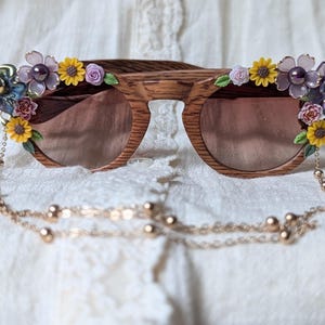 Könnte beinhalten: Holz Sonnenbrille mit Blumenmuster. Die Sonnenbrille hat einen braunen Rahmen und braune Gläser. Die Blumen sind aus Stoff und in den Farben Rosa, Lila, Gelb und Blau gehalten. Die Sonnenbrille hat eine goldene Kette daran.