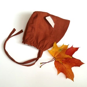 Puede incluir: Un gorro de bebé de lino marrón con una solapa de oreja de tela blanca y un cierre de corbata. El gorro está sobre una superficie blanca con dos hojas de otoño.