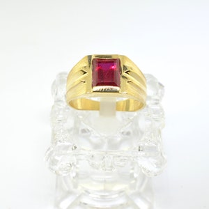 May include: A gold ring with a red gemstone. The ring has a textured band with a rectangular shape.