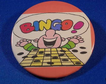 May include: A pink and white round button featuring a cartoon character playing bingo. The character is a pink person with a big smile and a blue hat. The word "Bingo!" is written in rainbow colours above the character.