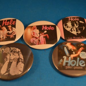 May include: Five round pinback buttons with different images of the band Hole. The buttons feature the band's name and album titles, including "Live Through This" and "Celebrity Skin".