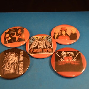 May include: A collection of five round, orange-colored pinback buttons. Each button features a different design, including band photos and graphics. The text "THE SMASHING PUMPKINS" is visible on one button.