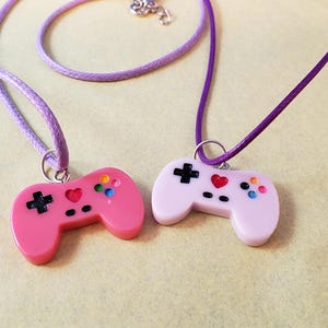 May include: Two pendant necklaces with game controller charms. One charm is pink, the other is light pink. Both have a purple cord. The controllers have black directional pads, colorful buttons, and a red heart.