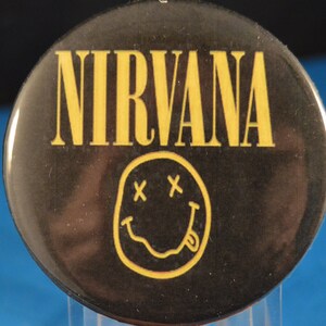 May include: A black and yellow button with the word "NIRVANA" in yellow letters and a yellow smiley face with an "X" for eyes.