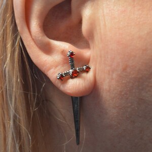 May include: A black dagger earring with red gemstones. The earring is worn in the upper lobe of the ear.