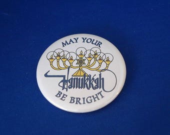 May include: White round button with the text "May Your Hanukkah Be Bright" in blue lettering. The button features a stylized image of a menorah with nine candles, a Star of David, and a blue and white background.