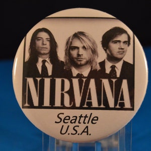 May include: Black and white photo of the band Nirvana. The band members are wearing suits and ties. The band's name is written in large, bold letters below the photo. The text "Seattle U.S.A." is written below the band's name.