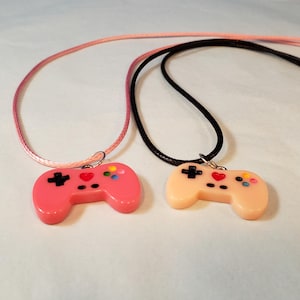 May include: Two pendant necklaces featuring game controller charms. One charm is pink, the other is peach. Each controller has a black cross, a red heart, and colorful buttons. The necklaces have pink and black cords.
