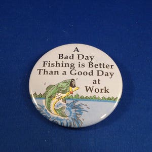 May include: A white button with a cartoon fish jumping out of the water. The button says "A Bad Day Fishing is Better Than a Good Day at Work".