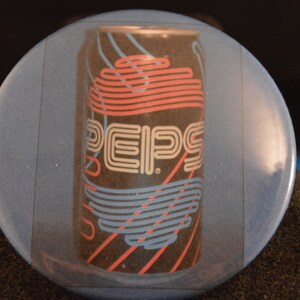 May include: A round, blue button featuring a retro design of a Pepsi can. The can is black with red, white, and blue lines and the Pepsi logo. The button has a glossy finish and is set against a dark background.