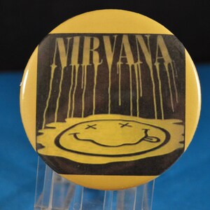 May include: Yellow round button with a black and white image of the band Nirvana's logo. The logo is a smiley face with an 'X' for eyes and a line for a mouth. The word 'Nirvana' is printed above the smiley face.
