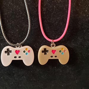 May include: Two pendant necklaces featuring game controller designs. One controller is white with a silver chain, the other is tan with a pink cord. Both controllers have a black cross, a red heart, and colorful buttons.