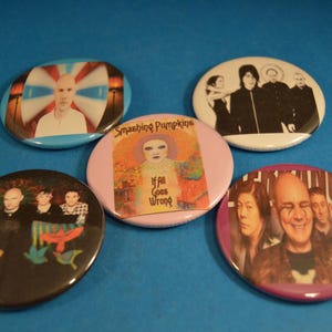 May include: Five round button pins featuring various music artists. The pins are colorful and feature images of the artists, including Billy Corgan of The Smashing Pumpkins, Morrissey, and The Cure.