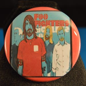 May include: A round button with a red border featuring the band Foo Fighters. The image inside shows the band members in a stylized illustration against a light blue background. The band's name is printed at the top.