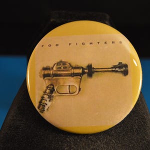 May include: A yellow round button with the text "Foo Fighters" and a black and white illustration of a futuristic gun.