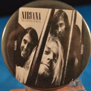 May include: A black and white button featuring a photo of the band Nirvana. The button has the band's name "NIRVANA" and the album title "THE CHOSEN REJECTS" printed in white text.