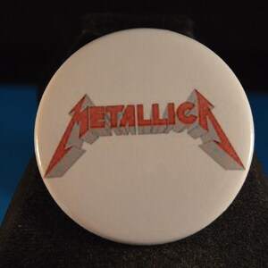 May include: White button with the band name "Metallica" in red and gray, stylized with a lightning bolt design. The button is round and has a glossy finish, perfect for fans of the heavy metal band.