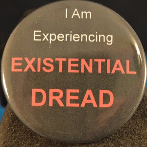 May include: A black button with white text that reads "I am Experiencing EXISTENTIAL DREAD" in red letters.