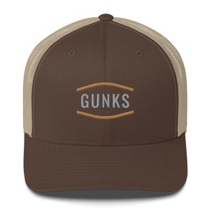 May include: A brown and tan trucker hat with a brown brim and a tan mesh back. The hat has an embroidered logo that says "GUNKS" in a light blue color with a brown outline.
