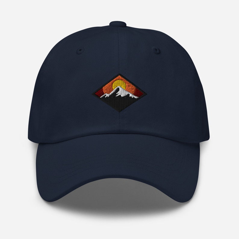 May include: A navy blue baseball cap with an embroidered patch featuring a mountain range with a setting sun. The patch is in a diamond shape with orange, yellow, and red colors.