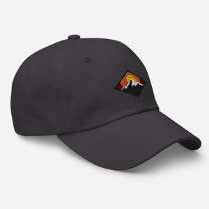 May include: A dark gray baseball cap with an embroidered patch featuring a mountain range and a setting sun. The patch is in a diamond shape with orange, red, and white accents.