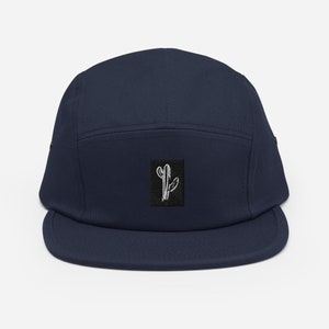 May include: A navy blue 5-panel cap with a black square patch featuring a white line drawing of a cactus.