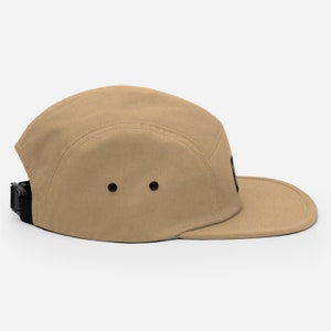 May include: A tan 5-panel cap with a black strap and a black patch on the side.