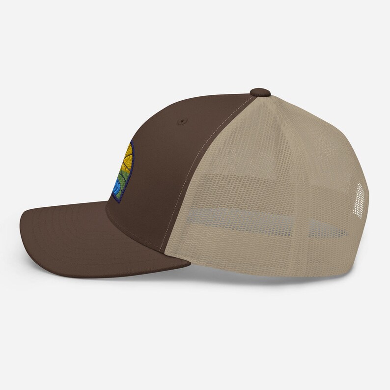 May include: A brown and beige trucker hat with a colorful embroidered patch on the front. The patch features a yellow sun, blue water, and green trees.