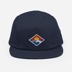Pode incluir: Um chapéu de 5 painéis azul marinho com um remendo branco em forma de diamante que apresenta uma cordilheira de montanhas estilizada e um sol amarelo, representando a bandeira do estado do Colorado.