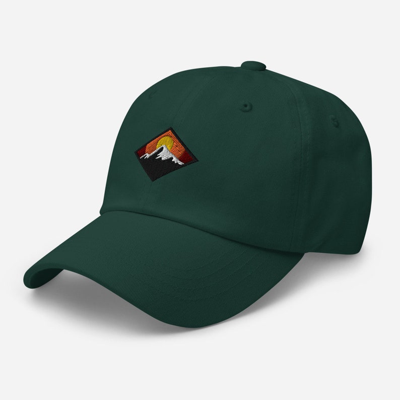 May include: A dark green baseball cap with an embroidered patch of a mountain range with a setting sun. The patch is in a diamond shape with orange, yellow, and white accents.