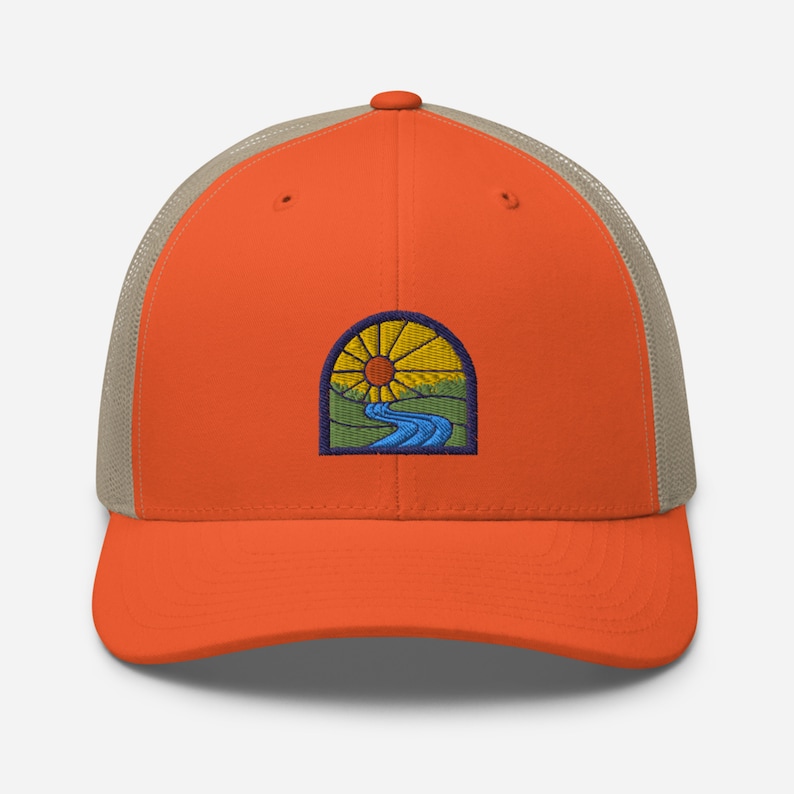 May include: Orange and beige trucker hat with an embroidered patch of a sun, river, and green hills in a stained glass style.