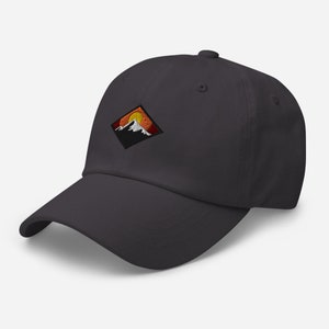 May include: A dark gray baseball cap with an embroidered patch of a mountain range with a setting sun. The patch is a diamond shape with orange, yellow, and white colors.