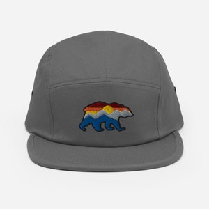 May include: A gray 5-panel cap with an embroidered design of a bear silhouette with a sunset and mountain range inside the bear.