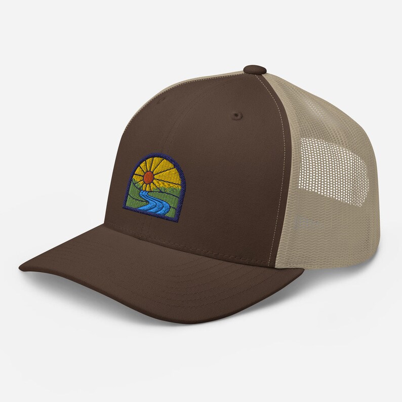May include: A brown and beige trucker hat with a patch of an embroidered sun, river, and hills.